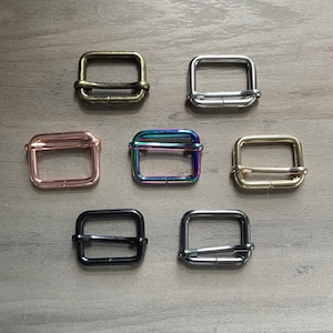 May include: Eight metal square slider buckles in different finishes: antique brass, silver, rose gold, iridescent, gold, black, and two shades of silver. The buckles are all the same size and shape.