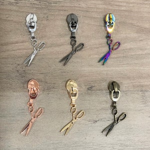 May include: Six different zipper pulls with a pair of scissors attached to each. The zipper pulls are in silver, black, rainbow, rose gold, gold, and bronze.