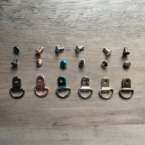 May include: Assortment of metal D-rings in various finishes, including black, rose gold, iridescent, silver, and brass. The D-rings are arranged in a row on a wooden surface.