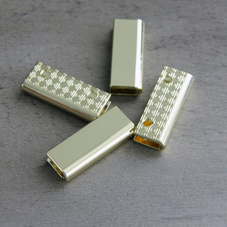 May include: Four gold-colored metal slider buckles with a textured pattern. The buckles are rectangular and have a hole on each end.