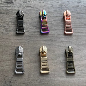 Op de afbeelding: Zes ritsen in verschillende kleuren, waaronder zwart, zilver, goud, rosé goud en regenboogkleuren. Elke rits heeft een rechthoekige vorm met vier gaten.