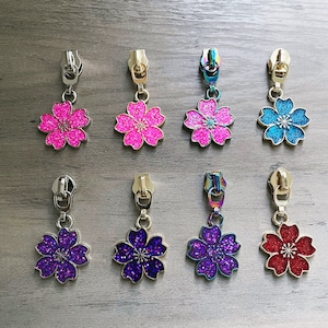 May include: Ten zipper pulls with a flower design. The flowers are pink, blue, purple, and red. Each zipper pull has a metal loop at the top.