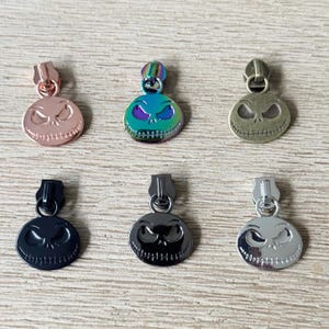 May include: Six metal zipper pulls in various colours, each featuring a circular design with a carved face. Colours include rose gold, iridescent, antique brass, black, gunmetal, and silver. The pulls are designed for use on clothing or accessories.