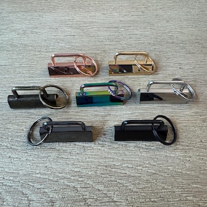 May include: A collection of seven metal purse clasps in various finishes, including rose gold, gold, iridescent, silver, gunmetal, and black. Each clasp features a rectangular base with a handle and a circular ring.