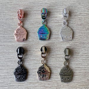 May include: Six different zipper pulls with cupcake charms. The charms are in various colors, including silver, gold, black, and rainbow. The zipper pulls are all metal.