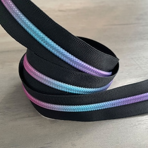May include: A coiled length of black zipper tape with a gradient of colors. The zipper teeth are a blend of pink, blue, and purple, creating a colorful accent against the black fabric. The zipper is likely for sewing or crafting projects.