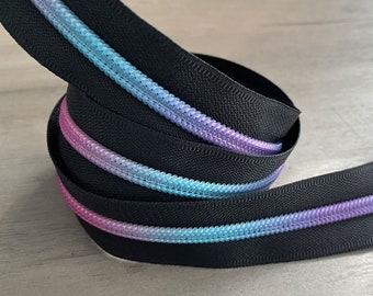 Puede incluir: Una cinta de cremallera negra enrollada con un degradado de colores. Los dientes de la cremallera presentan una mezcla de rosa, azul y morado, creando un acento colorido sobre la tela negra. Adecuado para proyectos de costura o manualidades.