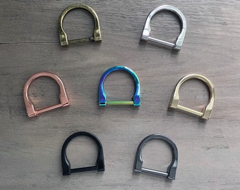 Può includere: Sette anelli a D in metallo di diversi colori. Gli anelli sono disposti in cerchio su una superficie di legno. I colori sono oro, argento, oro rosa, blu iridescente, oro, nero e canna di fucile.