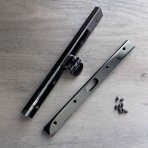 May include: Two black metal brackets with multiple holes and a small black plastic piece with a metal spring. A pile of small black screws are on a wood surface.