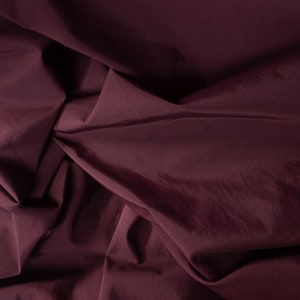 Puede incluir: Primer plano de una tela de color burdeos oscuro con una textura suave y sedosa. La tela est&aacute; doblada y drapeada, creando l&iacute;neas suaves y fluidas.