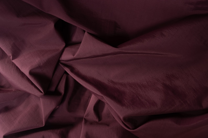 Puede incluir: Primer plano de una tela de color burdeos oscuro con una textura suave y sedosa. La tela est&aacute; doblada y drapeada, creando l&iacute;neas suaves y fluidas.