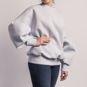 Puede incluir: Una sudadera de color azul claro con mangas largas y un corte holgado. Las mangas tienen un aspecto abullonado y fruncido.