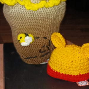 May include: A handmade crochet honey pot with a yellow ruffled trim and a small bee applique. Next to it is a yellow and red crochet hat. The honey pot has a brown and tan color gradient.