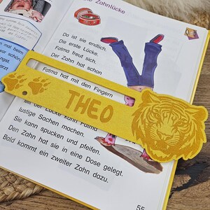 May include: A yellow bookmark with the name "THEO" and a tiger head design. The bookmark is placed on an open book with German text and illustrations. The bookmark also features paw prints.