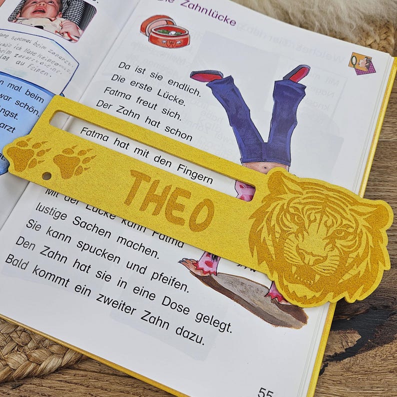 May include: A yellow bookmark with the name "THEO" and a tiger head design. The bookmark is placed on an open book with German text and illustrations. The bookmark also features paw prints.