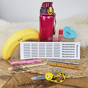 Könnte beinhalten: Ein Set mit Schulbedarf, bestehend aus einer magentafarbenen Trinkflasche, einem gelben bananenförmigen Etui, einem Klebestift, einem herzförmigen Behälter, einem Bogen Aufkleber mit dem Text 'Wunschname', einem rosa Stift, zwei Pinseln, einem Bleistift, einem Filzstift und einer bienenförmigen Schere.