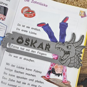 May include: A gray dragon-shaped bookmark with the name "OSKAR" printed on it, resting on the pages of a children's book. The book is open, displaying colorful illustrations and German text.