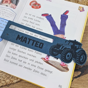May include: A navy blue bookmark with the name "MATTEO" and a tractor design. The bookmark is placed on an open book with German text and illustrations. The page number "55" is visible.
