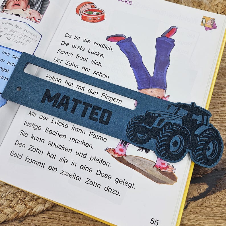 May include: A navy blue bookmark with the name "MATTEO" and a tractor design. The bookmark is placed on an open book with German text and illustrations. The page number "55" is visible.