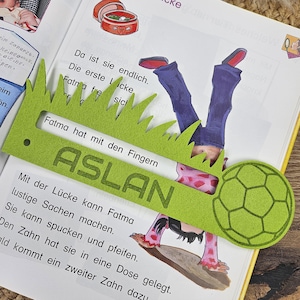 May include: A green felt bookmark with a grass-like edge and the name "ASLAN" printed on it. The bookmark is placed over an open book with illustrations and text. A soccer ball shape is attached to the bookmark.