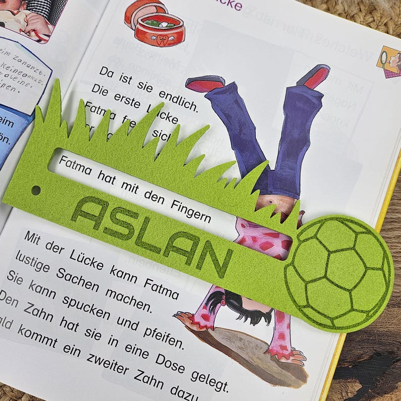 May include: A green felt bookmark with a grass-like edge and the name "ASLAN" printed on it. The bookmark is placed over an open book with illustrations and text. A soccer ball shape is attached to the bookmark.
