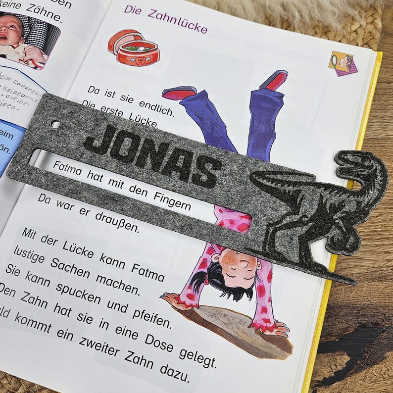 May include: A gray felt bookmark with the name "JONAS" in large black letters. A black dinosaur silhouette is on the right side. The bookmark is placed on an open book with colorful illustrations and German text.