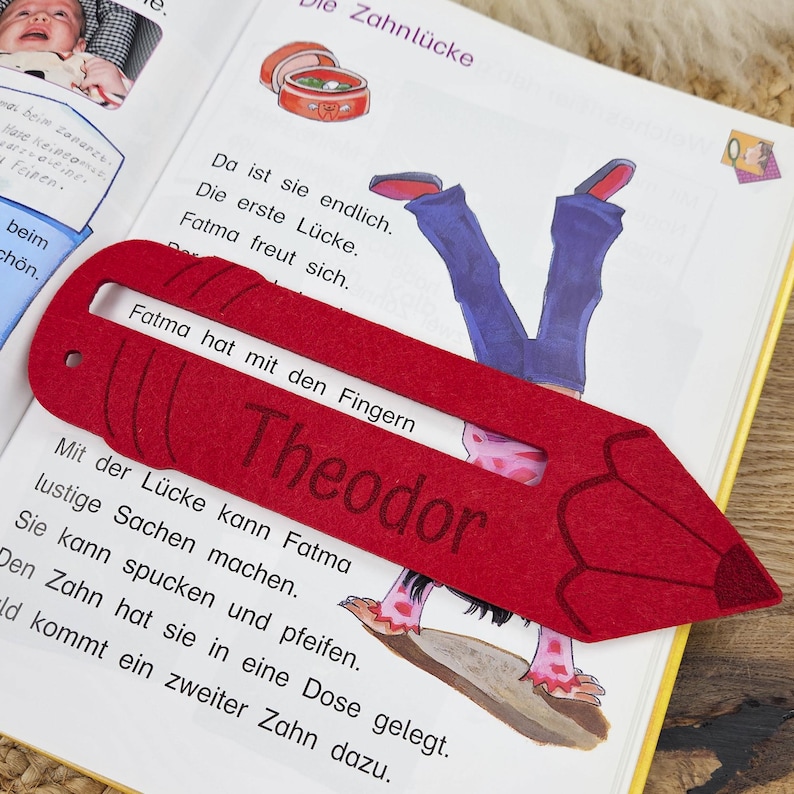 May include: A red felt pencil-shaped bookmark with the name "Theodor" printed on it, resting on an open book. The book's pages feature German text and illustrations. The bookmark is designed to mark a page in a book.