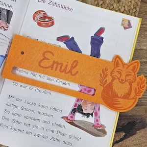 May include: Orange felt bookmark with a fox design and the name "Emil". The bookmark is placed over an open book with illustrations and German text. The text "Fatma hat mit den Fingern" is visible.