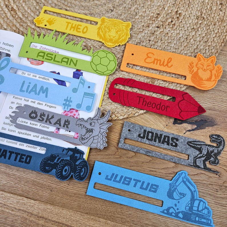 May include: A collection of colorful felt bookmarks with names and illustrations. The bookmarks feature names like Theo, Aslan, Liam, Oskar, Emil, Theodor, Jonas, and Justus, along with images of a tiger, soccer ball, musical notes, dragon, fox, pencil, and dinosaur.