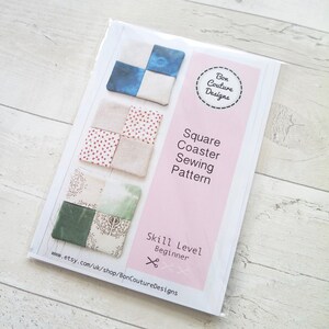 May include: A pink and white card with a sewing pattern for square coasters. The card shows three different designs for the coasters, each with a different fabric pattern. The card also includes the text "Square Coaster Sewing Pattern" and "Skill Level Beginner".