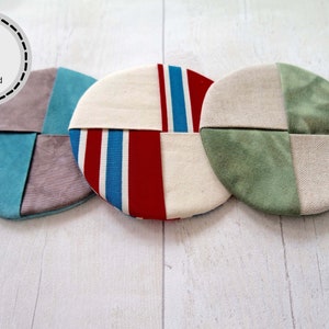 May include: Three round fabric coasters with different patterns. The coasters are made of a patchwork of fabric squares. The patterns include a blue and gray pattern, a red and blue striped pattern, and a green and white tie-dye pattern.