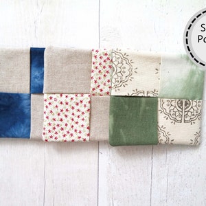 May include: A patchwork quilt block made of nine squares of fabric in various colors and patterns, including beige, blue, green, and white. The fabric squares are sewn together to create a geometric design.