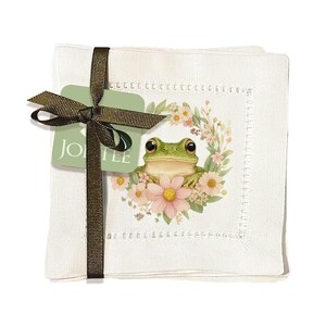 May include: A set of white linen coasters tied with a brown ribbon and a tag that reads "JOLEE". Each coaster features a green frog illustration surrounded by a floral wreath with pink and white flowers.