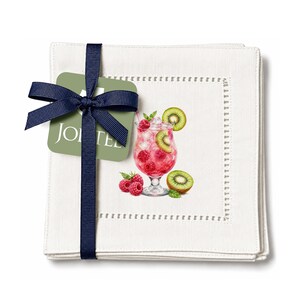 Peut inclure: Une pile de serviettes en tissu blanc carré avec un bord à jour, nouées par un ruban bleu marine et une étiquette verte marquée JOI TEE. La serviette supérieure présente une illustration imprimée d'un cocktail rose avec des framboises et du kiwi.