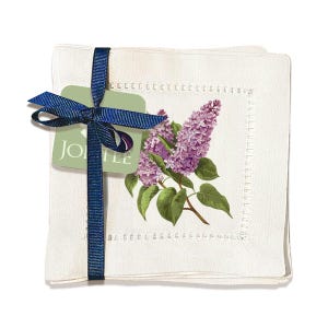 May include: Set of four white linen cocktail napkins with a lilac floral design and a blue ribbon. The napkins are folded and tied together with a blue ribbon and a green tag that says "JOLLEE".