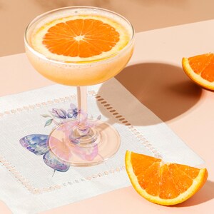 K&ouml;nnte beinhalten: Ein Glas Orangensaft mit einer Orangenscheibe als Garnitur, das auf einer wei&szlig;en Serviette mit einem Schmetterlingsmuster liegt. Zwei Orangenscheiben sind ebenfalls auf dem Tisch zu sehen.