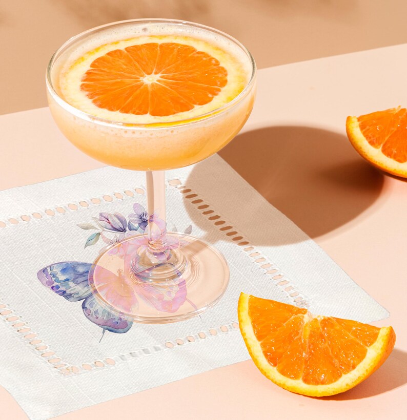 K&ouml;nnte beinhalten: Ein Glas Orangensaft mit einer Orangenscheibe als Garnitur, das auf einer wei&szlig;en Serviette mit einem Schmetterlingsmuster liegt. Zwei Orangenscheiben sind ebenfalls auf dem Tisch zu sehen.
