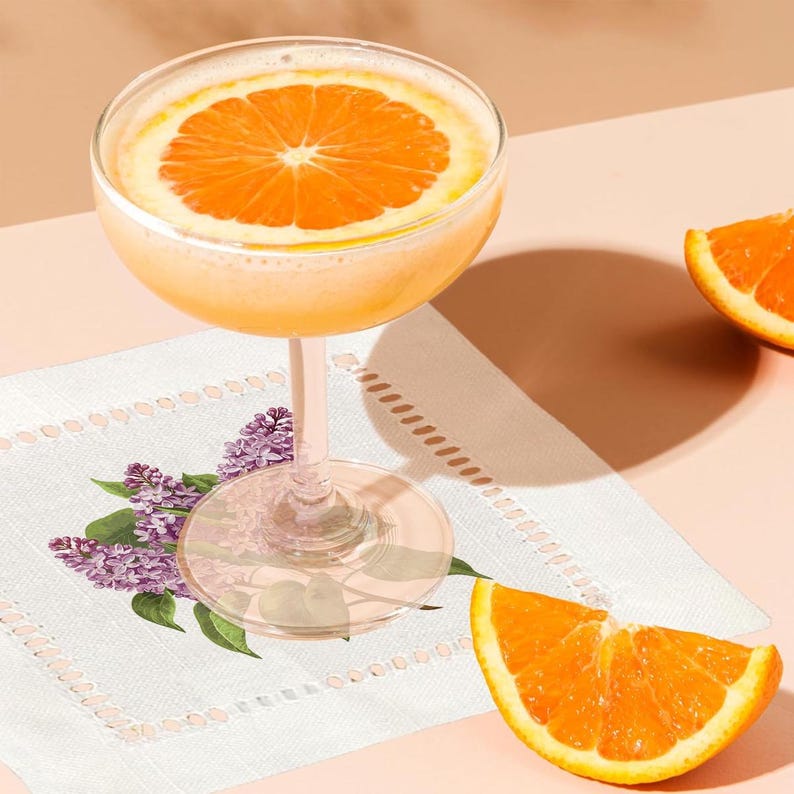 May include: A cocktail with a slice of orange in a coupe glass on a white linen napkin with a floral design. The napkin has a scalloped edge.