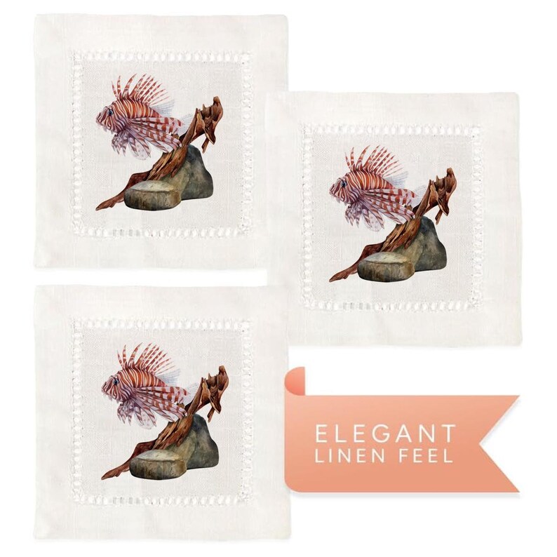 May include: Set of three linen cocktail napkins featuring a watercolor illustration of a lionfish perched on a piece of driftwood. The napkins have a white background with a delicate white embroidered border.