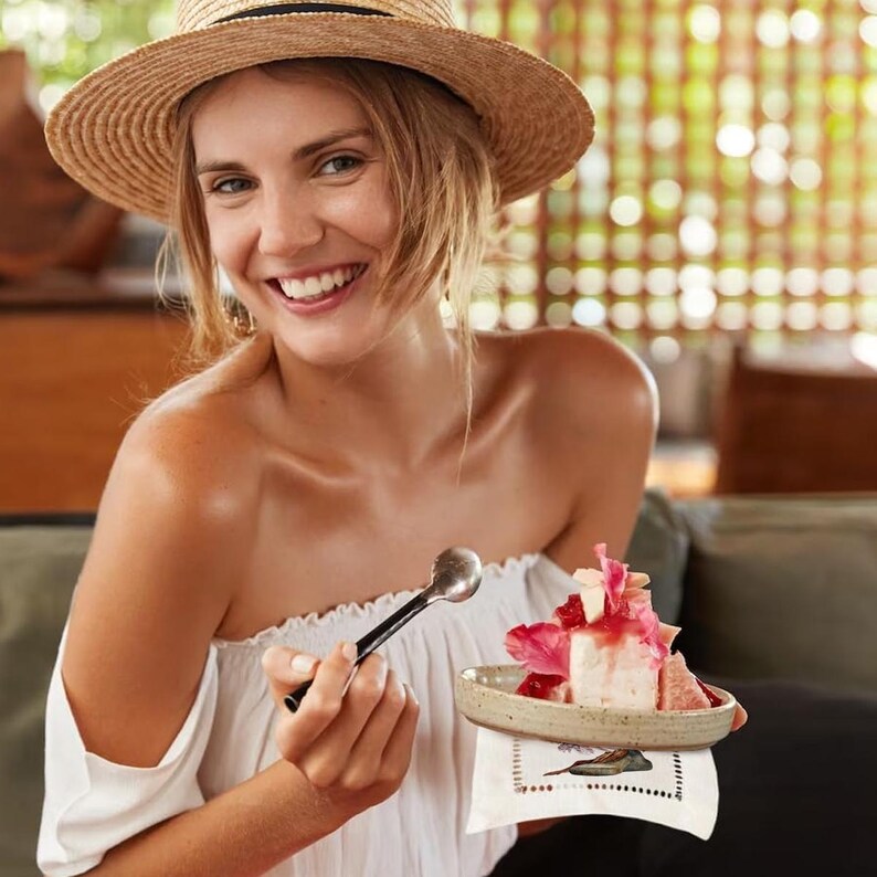 May include: A woman wearing a straw hat smiles while holding a spoon and a small plate with a pink and white dessert topped with fruit.