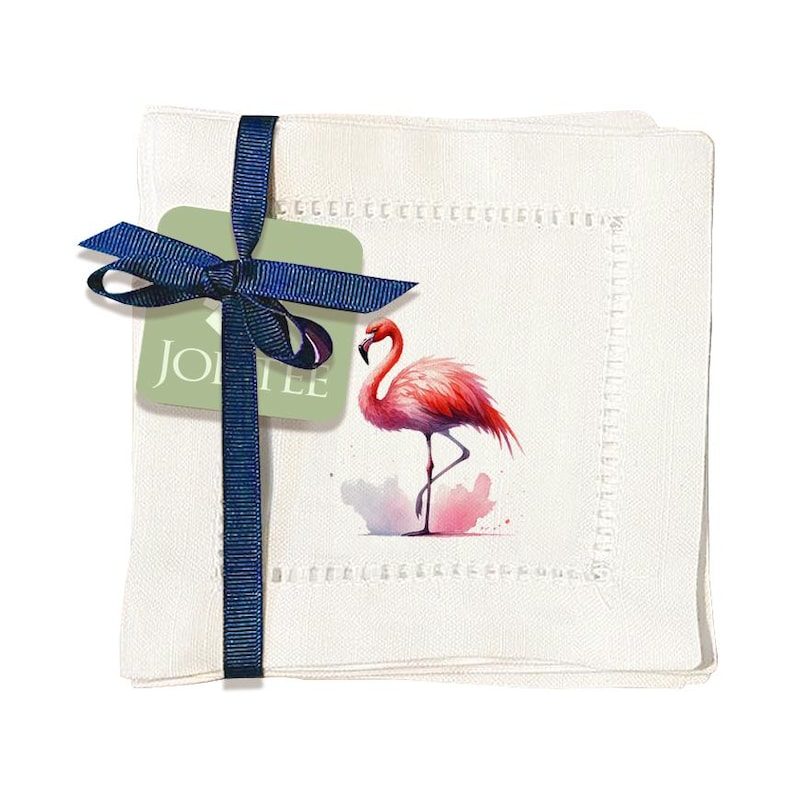 May include: Set of four white linen cocktail napkins with pink flamingo illustration and a blue ribbon tied around the stack. The napkins have a white stitched border and a green tag with the text "JOLTEE" on it.