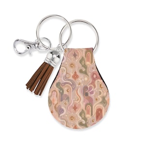May include: A teardrop-shaped keychain featuring a repeating pattern of arches, stars, and abstract shapes in muted pink, sage green, and cream tones. It includes a silver-toned metal split ring, a lobster clasp, and a brown faux leather tassel.