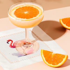 May include: A cocktail with a slice of orange in a coupe glass on a white napkin with a pink flamingo illustration.