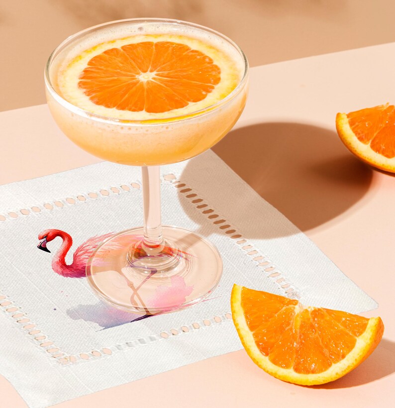 May include: A cocktail with a slice of orange in a coupe glass on a white napkin with a pink flamingo illustration.