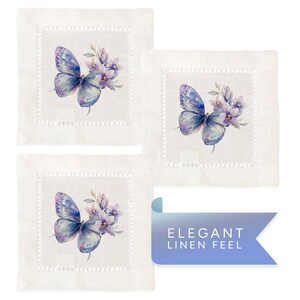 K&ouml;nnte beinhalten: Set aus drei wei&szlig;en Leinen-Servietten mit einer zarten, gestickten Bord&uuml;re. Jede Serviette zeigt eine Aquarell-Illustration eines lilafarbenen Schmetterlings mit Blumen.