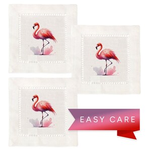 May include: Set of three white linen cocktail napkins with pink flamingo illustrations. Each napkin features a pink flamingo standing on one leg with a watercolor background. The napkins have a scalloped edge and the text "EASY CARE" is printed on a pink and purple ribbon.