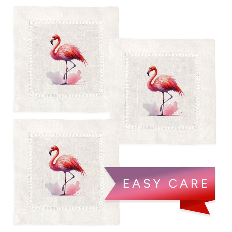 May include: Set of three white linen cocktail napkins with pink flamingo illustrations. Each napkin features a pink flamingo standing on one leg with a watercolor background. The napkins have a scalloped edge and the text "EASY CARE" is printed on a pink and purple ribbon.