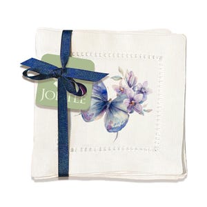 K&ouml;nnte beinhalten: Set aus vier wei&szlig;en Leinen-Cocktail-Servietten mit einem Design aus einem blauen Schmetterling und lila Blumen. Die Servietten haben einen zarten gestickten Rand. Die Servietten sind mit einem blauen Band und einem gr&uuml;nen Etikett mit der Aufschrift "JOLIE" zusammengebunden.