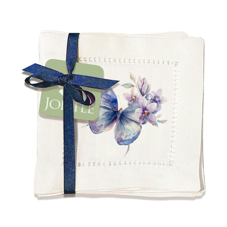 K&ouml;nnte beinhalten: Set aus vier wei&szlig;en Leinen-Cocktail-Servietten mit einem Design aus einem blauen Schmetterling und lila Blumen. Die Servietten haben einen zarten gestickten Rand. Die Servietten sind mit einem blauen Band und einem gr&uuml;nen Etikett mit der Aufschrift "JOLIE" zusammengebunden.