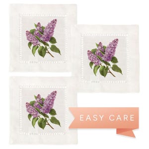 May include: Set of three white linen cocktail napkins with a lilac floral design. Each napkin features a cluster of purple lilacs with green leaves. The napkins have a scalloped edge and are machine washable.
